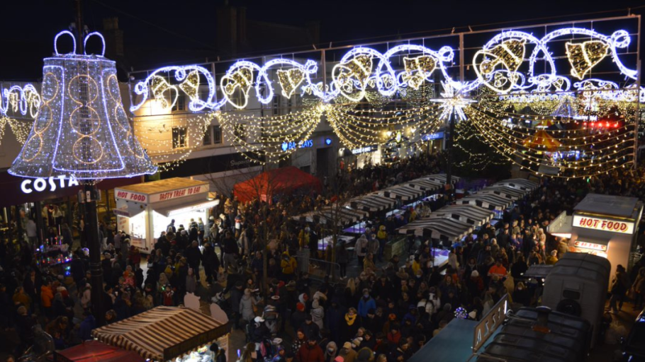 A packed town centre at night with bright Christmas lights, including a huge bell of lights above the crowds