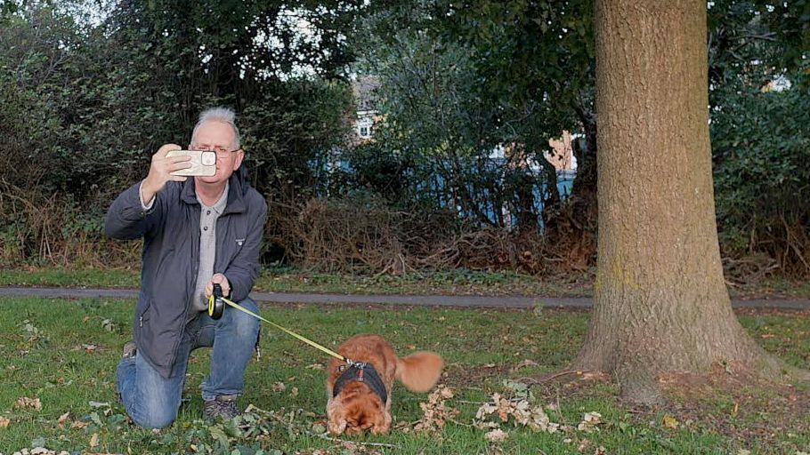 A man in a blue coat and blue jeans with a small brown dog next to a tree taking a photograph