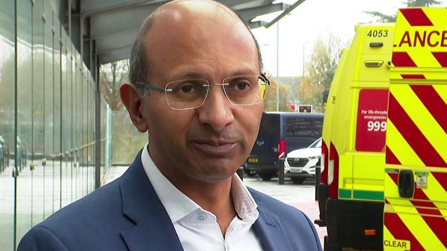Professor Kiran Patel stood in front of ambulances and wearing a suit and glasses.