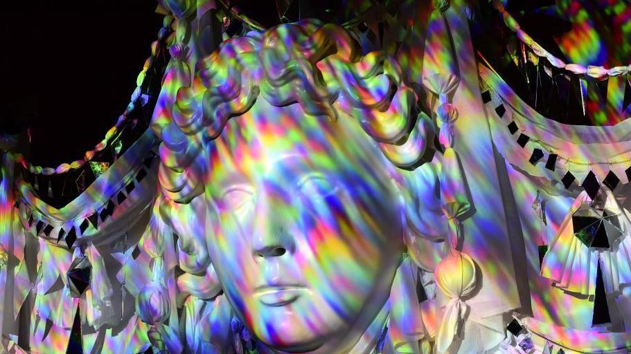A close-up of a face sculpture with multicoloured lights reflected on it.