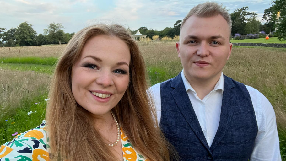 Woman with long hair in bright floral dress, with her husband who has short blonde hair and is wearing a waistcoat and shirt. They smile at the camera on a sunny day, standing in a field.