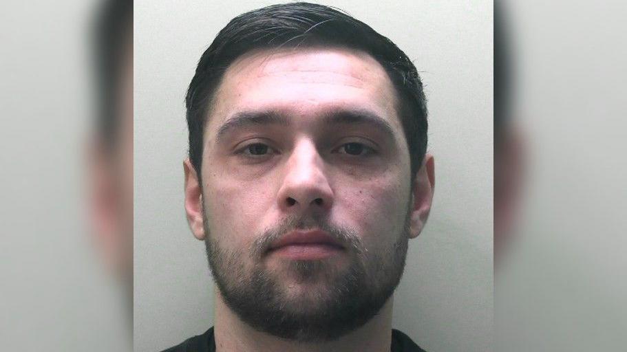 A man with short black hair and a scruffy black beard in a mug shot. He is wearing a black top, background of a cream wall. 