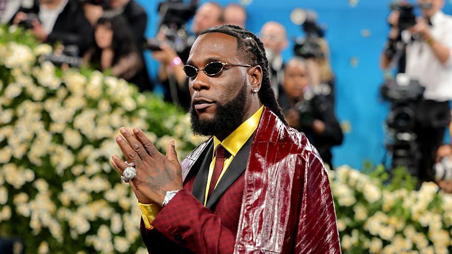 Burna Boy at the 2025 Met Gala in New York. He wears a red leather cape over a red suit with black lapels, a bright yellow shirt and red tie. He poses with his hands held together, a large diamond ring on his left little finger. Photographers can be seen in the background as he walks the red carpet.