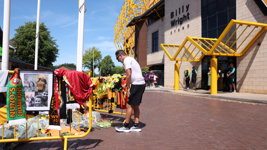 A man in a white top, black shorts and black and white trainers bends down by bunches of flowers and football scarves. Behind him is a football stadium, with a sign on the side saying the Billy Wright stand