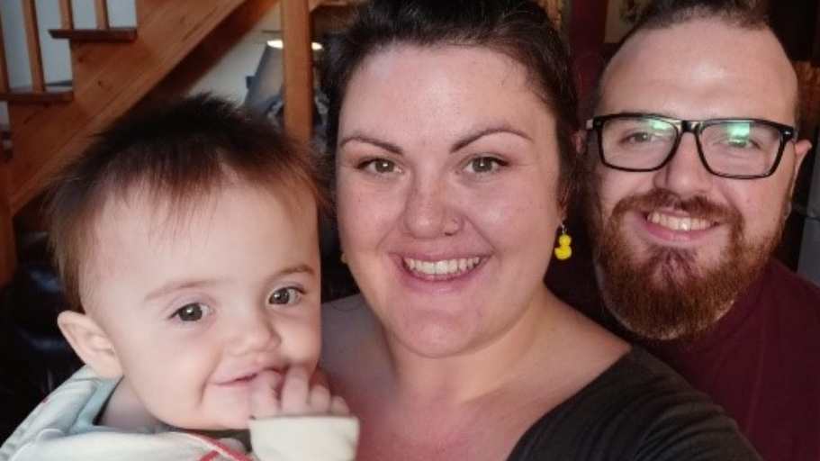 Rachel who had dark hair in a bun and rubber duck earrings in her ears, smiling and looking at the camera, with baby Eleanor in her arms and Chaz, who has glasses and a ginger beard, stood beside her.