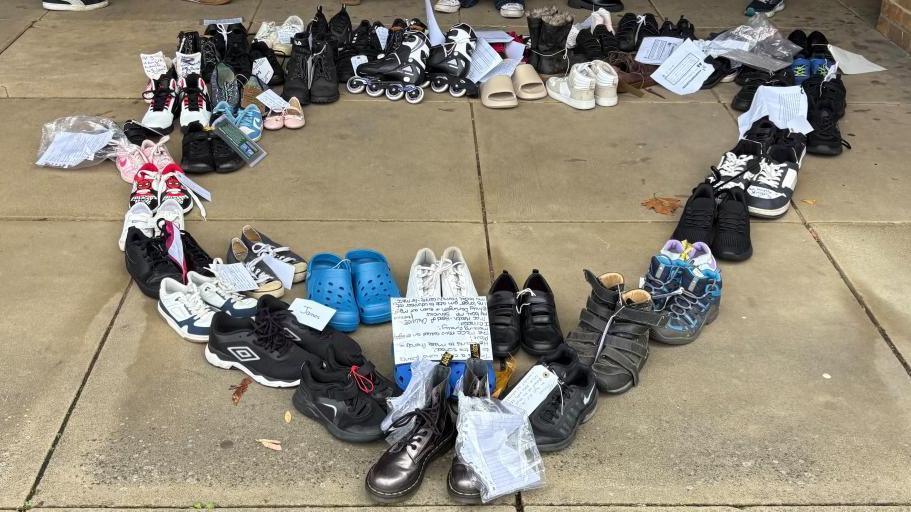 Shoes of various colours including black, blue and pink, arranged in pairs, some with white tags tied onto them with names or other messages. They are arranged in a heart shape on paving slabs. Some people's feet are visible along the top of the picture.