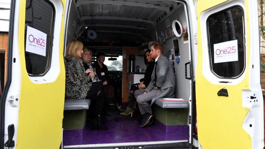 The Duke and Duchess of Sussex in the back of the One25 outreach van, which has yellow doors and purple floor, speaking to members of the charity team.