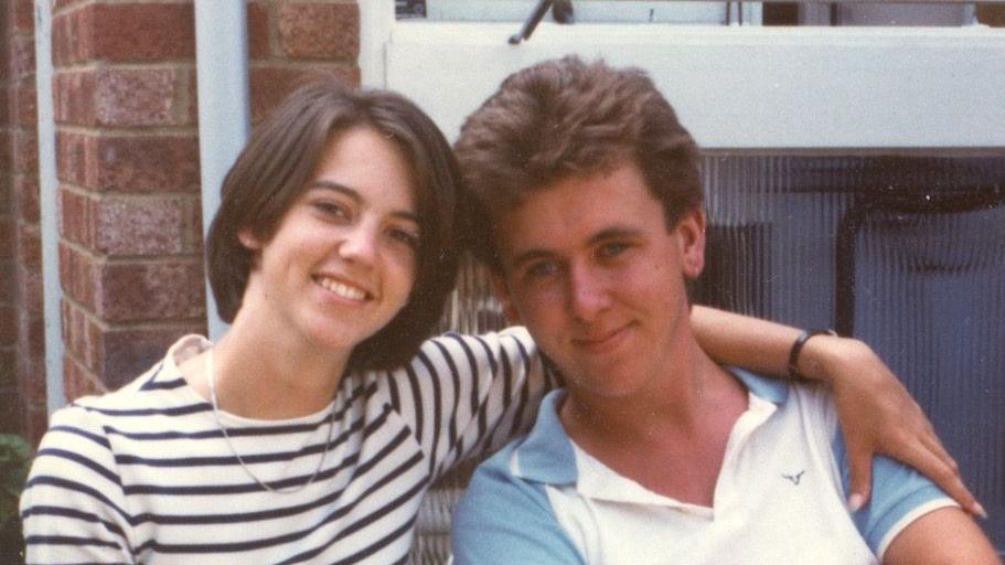 An old photo of Karen Goddard with her arm around her husband Tim. She has a brown bob and wears a white long sleeved top with black stripes and a necklace around her neck. Tim has brown hair and wears a white polo top with blue short sleeves.