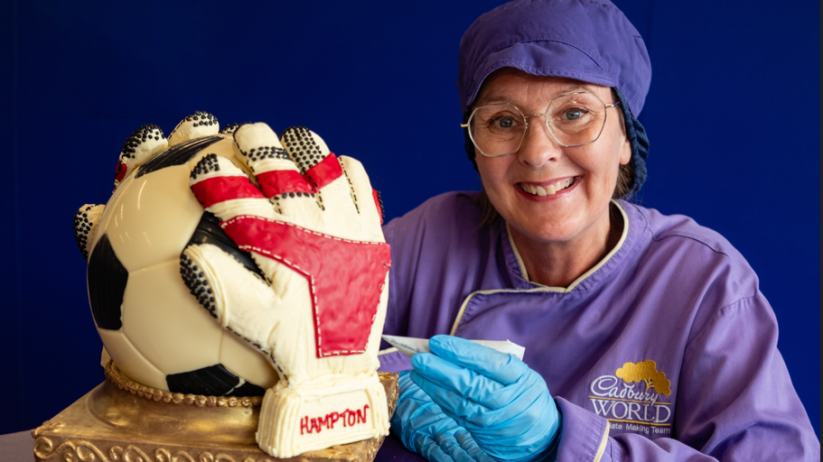 A woman sitting with a chocolate sculpture of a goalie. The sculpture is of a football with black and white panels and a pair of gloves wrapped around it. The gloves are white and red in colour.