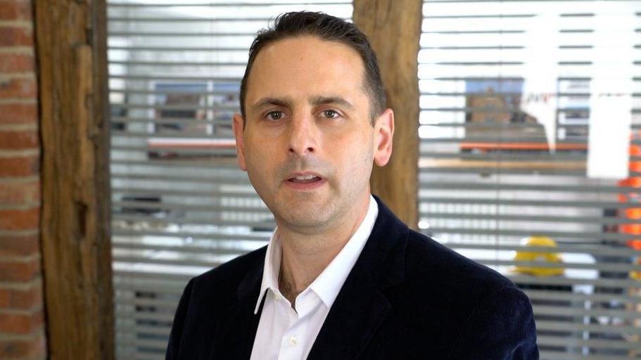 A man wearing a white shirt and a black jacket. He is looking directly into the camera. 