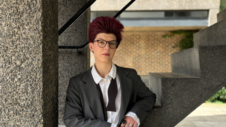 Zadirko is leaning against some concrete stairs with one arm resting on a step. They are wearing a grey blazer, white shirt and undone bow tie around their neck. Zadirko is also wearing glasses and has dark red hair that is spiked upwards.