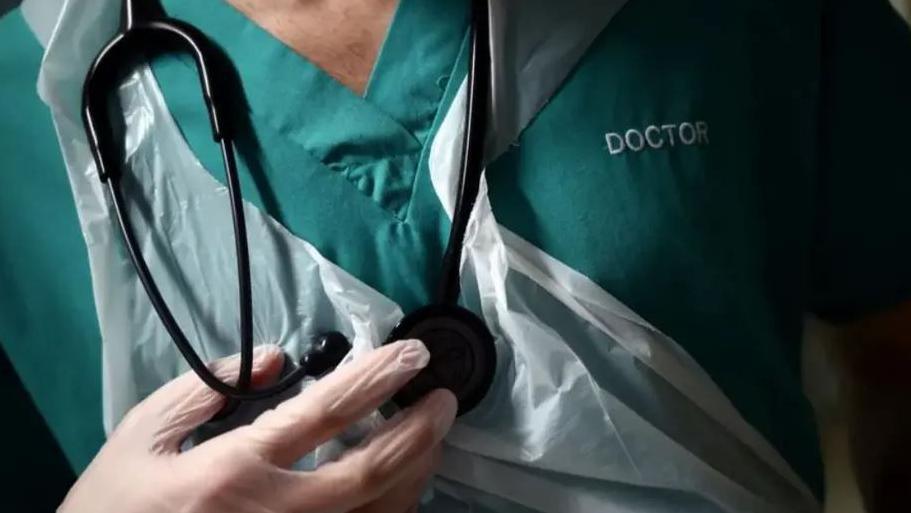 A close up of the chest of a doctor, who is wearing a green vest with the word doctor embroidered onto it. He is wearing white plastic gloves, and a white plastic apron and has a black stethoscope hanging around his neck.