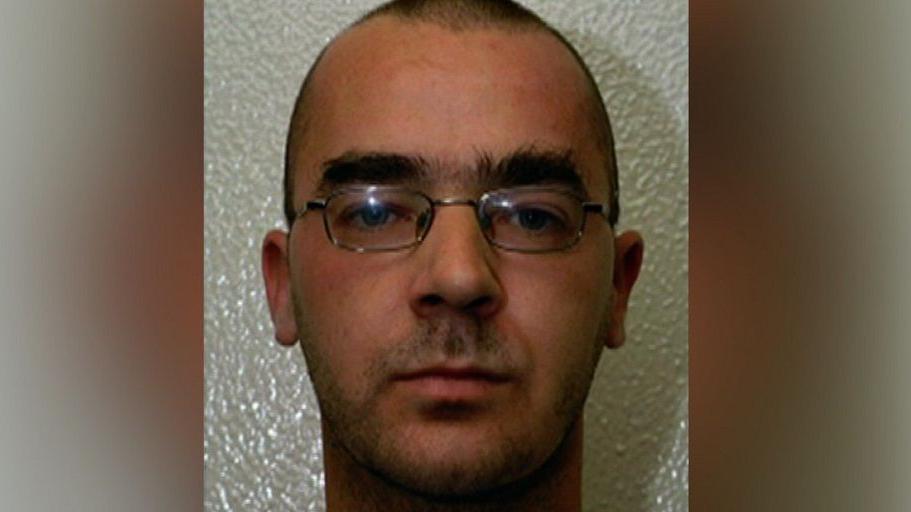 A man in his 20s, with close-cropped dark hair, thick eyebrows and metal-rimmed glasses, looking at the camera.