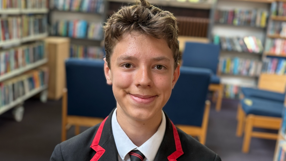 Daniel sitting in his school library. He has short brown hair, is wearing his black and red school uniform and is smiling.