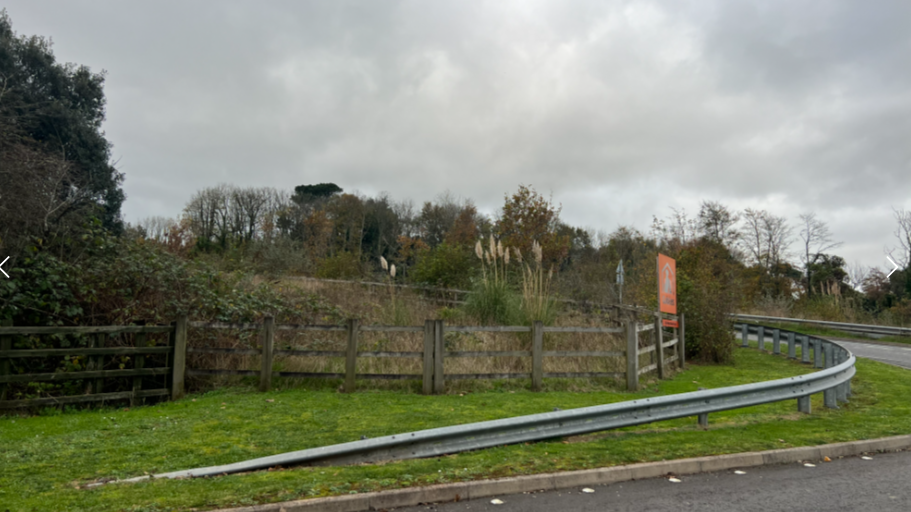 A grassy area near a road. On the right is the entrance to Gables in Plymouth. There is also a wooden fence which has bushes and trees behind it.