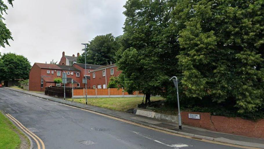 A street view image shows the road with trees to the right and red brick houses further at the top. Road markings such as double yellow lines can be seen at the start of the road.