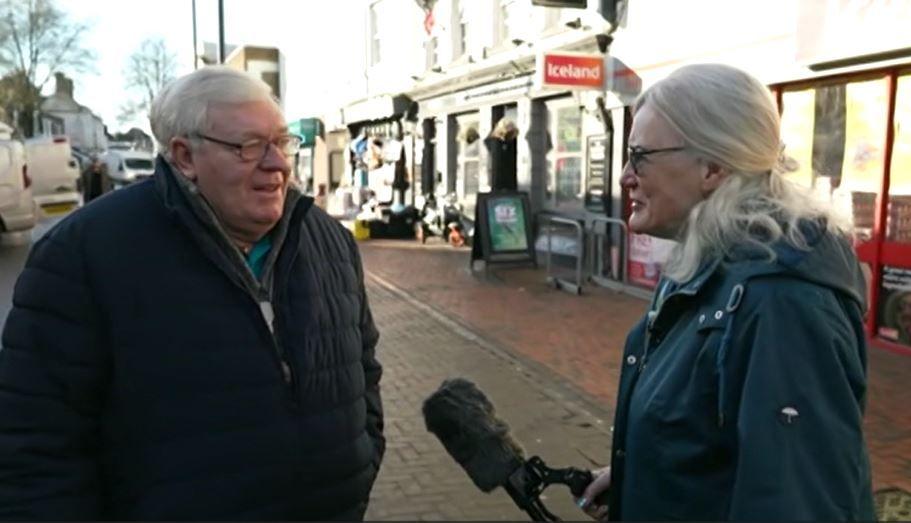 A man with short white hair and wearing metal rimmed glasses looks at Anita Stanley as she holds a microphone with a furry grey cover towards him. The man wears a black, padded coat. They stand on a high street with shops behind them.