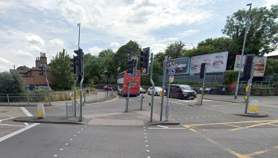 Traffic signals at junction of Station Road/A607 junction with  red and white bus passing billboards in the background