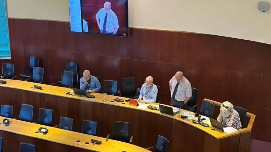 Pembrokeshire county council meeting with desks and chairs in tiers and four cabinet members in their seats, one standing up