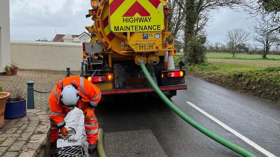 A man in an orange high vis jacket and trousers leaning down to clear a drain, with a highway maintenance van on the road.