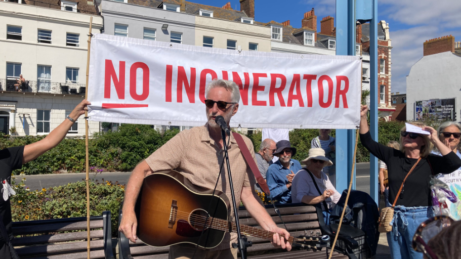Billy Bragg holding a guitar, wearing a beige shirt and dark sunglasses in front of a microphone.