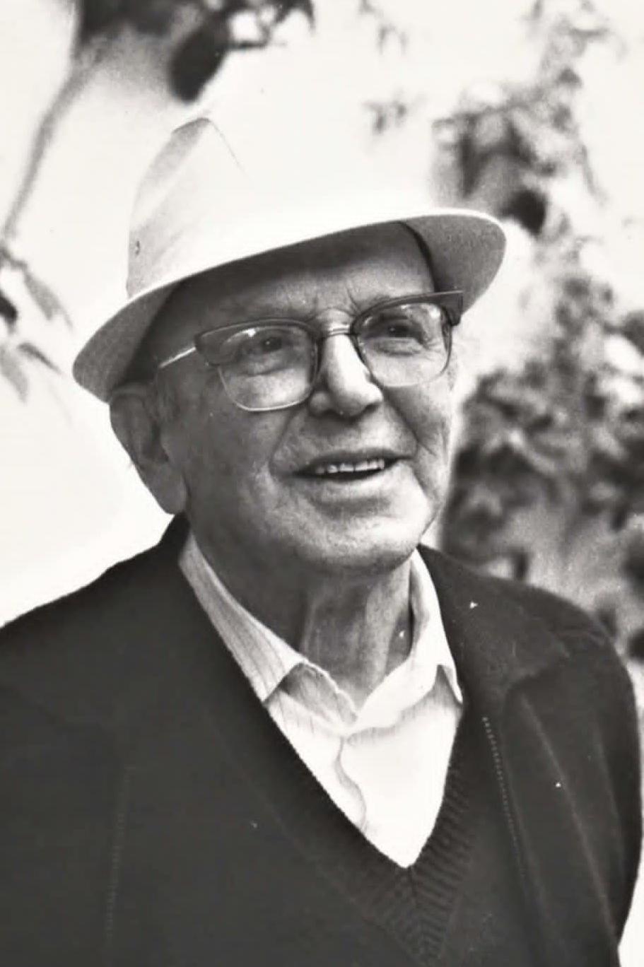 A black and white photograph of an elderly man in glasses and a white hat. He is wearing a shirt and a V-neck jumper. He is smiling and looking to the left of the camera. 