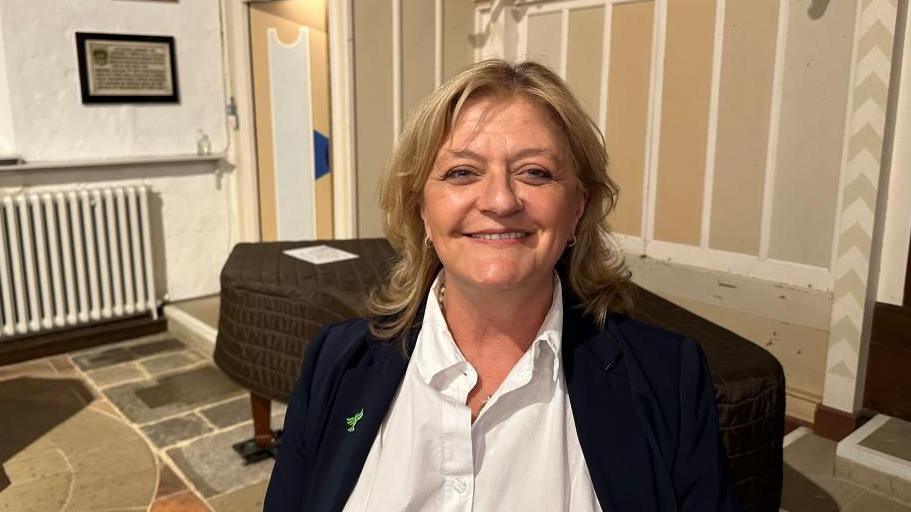 A woman is standing in a room which looks like an old town hall. She has medium length blonde hair and is smiling. She is waring a blue jacket and white top. 