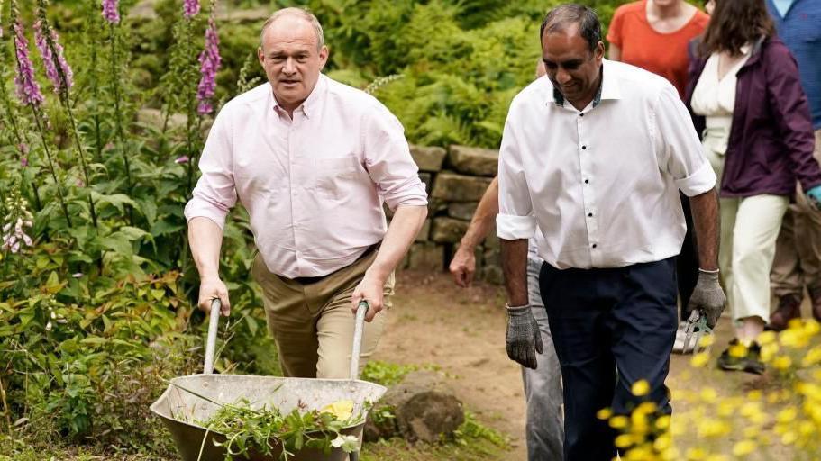 Ed Davey pushes a wheelbarrow containing plants in a garden, accompanied by Shaffaq Mohammed, with other people following. Both men are in shirtsleeves, while Mr Mohammed wears gardening gloves.