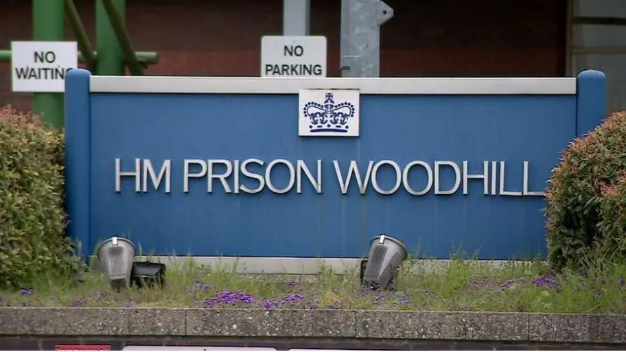 Entrance to HMP Woodhill, showing blue prison sign in the centre of some hedges.
