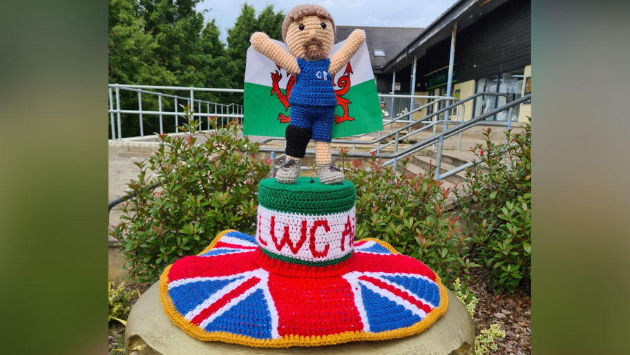 Knitted postbox topper of Paralympian Aled Sion Davies holding a Welsh flag and standing on a Union flag