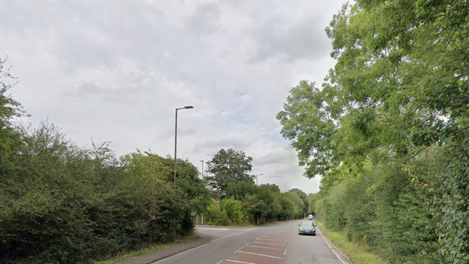 The A1000 road with the junction for the Oxlease Drive on the left side. There are tall hedges and trees lining the road, and a narrow pavement on the left side. There is a turning area marked in red with white chevrons in the middle of the road.