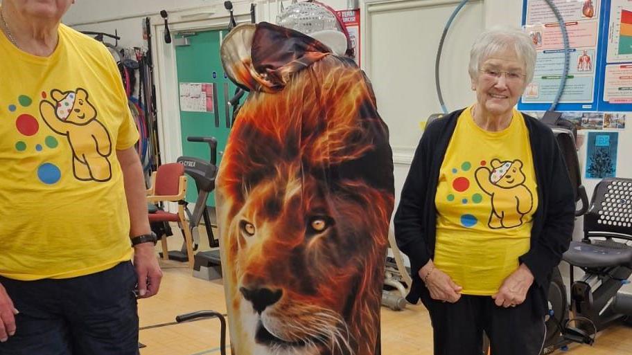 A woman in a fancy dress outift that has a lion on it standing in between two people in yellow t-shirts. They are standing in a gym in front of weight machines.