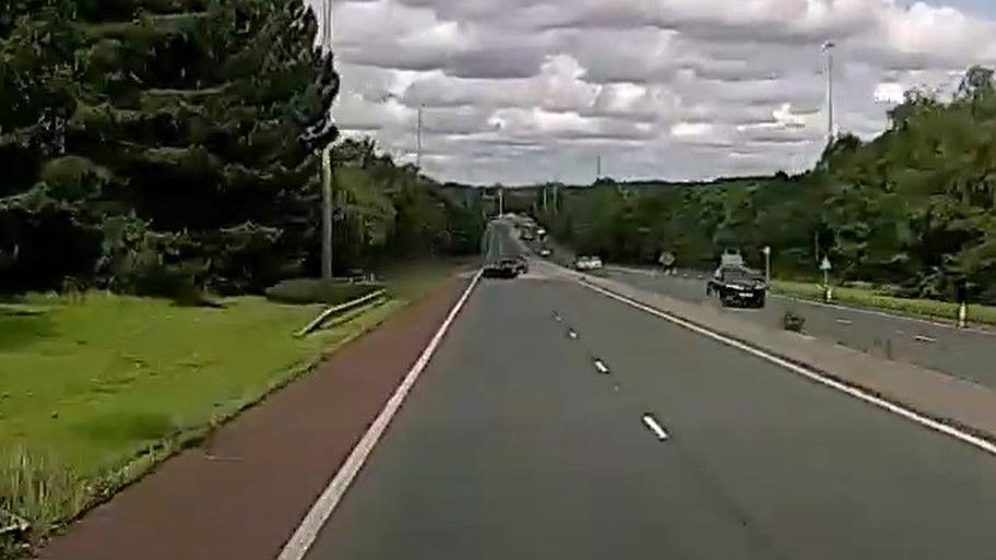 A still image showing a grey Ferrari car on a dual carriageway swerving left towards a slip road, where the police have blurred out the victim's vehicle.