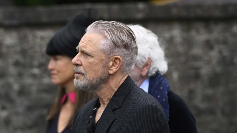 Adam Clayton, with grey hair and grey beard, wearing a dark suit and dark shirt, at the funeral of Lord Mount Charles.