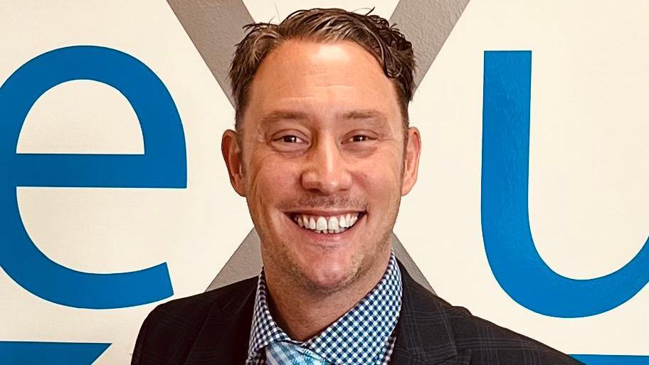 A man with short dark hair is wearing a black suit jacket, blue checked shirt and blue checked tie and is smiling. He is standing in front of a white wall with blue lettering.