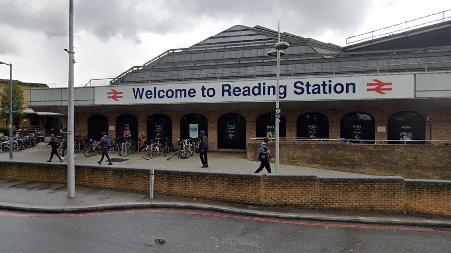 A sign on a single-storey brick building with a pyramid-shaped roof reads "Welcome to Reading Station". Bike racks and pedestrians are in front of the building.