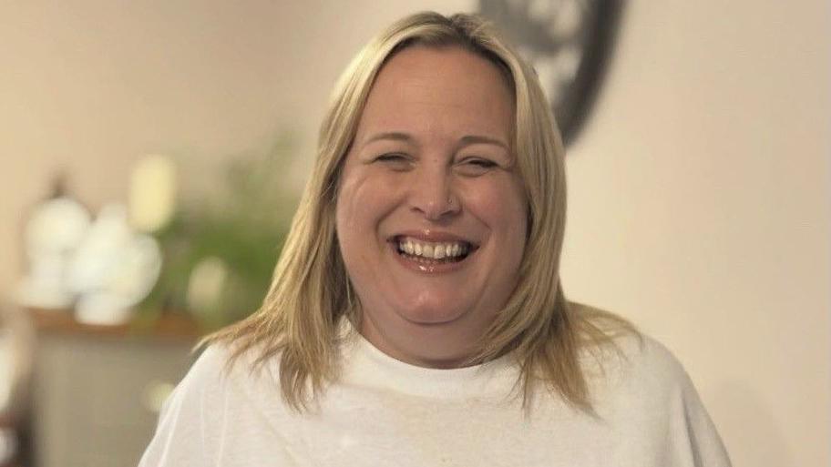 A woman is standing indoors, smiling widely at the camera and wearing a white T-shirt. The background is softly blurred, with household items and greenery, such as a clock and plants. She has blonde hair just past her shoulders.