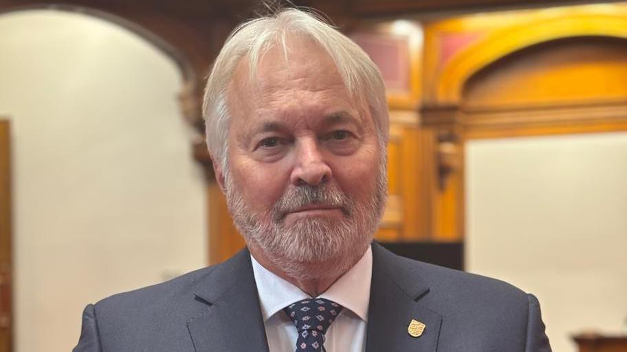 A man wearing a suit and tie, only shoulders up in vision, standing in the assembly chamber blurred in the background.