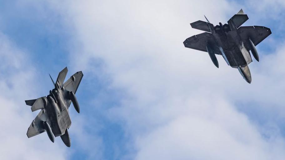 Two grey coloured American fighter aircraft in flight. Their undersides are visible and they have what appear to be aviation fuel drop tanks beneath their wings.