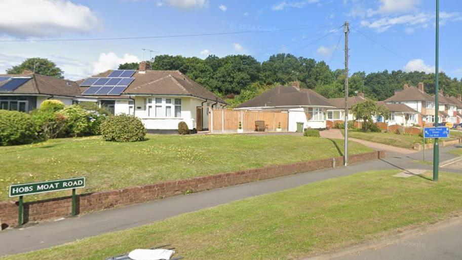 A sign for Hobs Moat Road can be seen on the pavement of a grassy verge. A umber of bungalows and two storey homes are pictured in the distance, with a grass lawn behind the road sign.