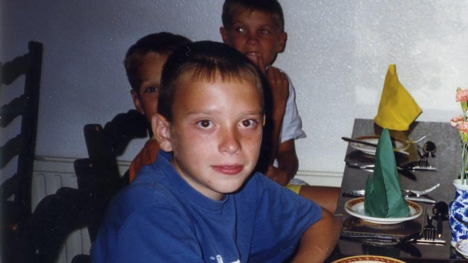 Anton wearing a blue t shirt on the left. There are several other people smiling at the camera as they sit down at a dinner table. There are green and yellow napkins on plates.
