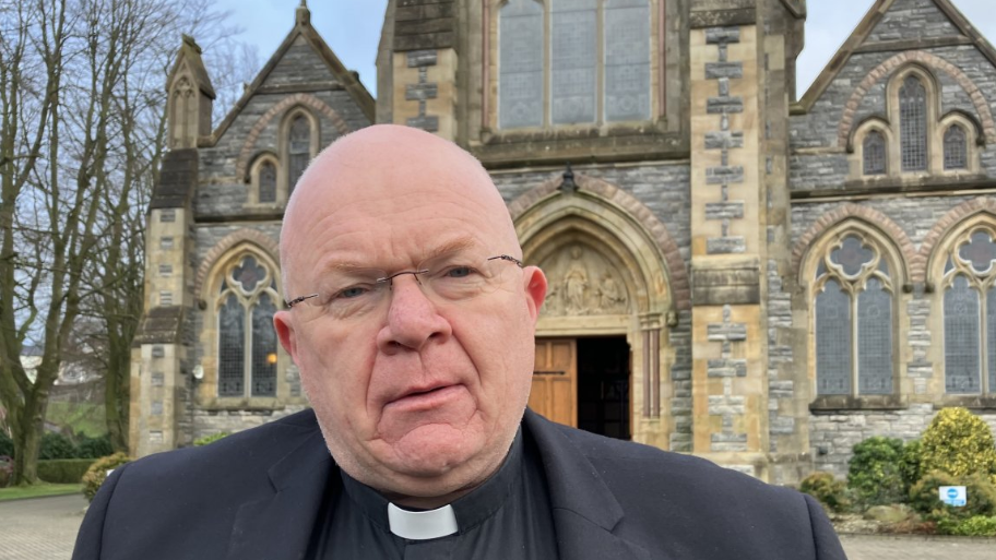 Shows Fr Michael McCaughey in a priests outfit standing in front of a church