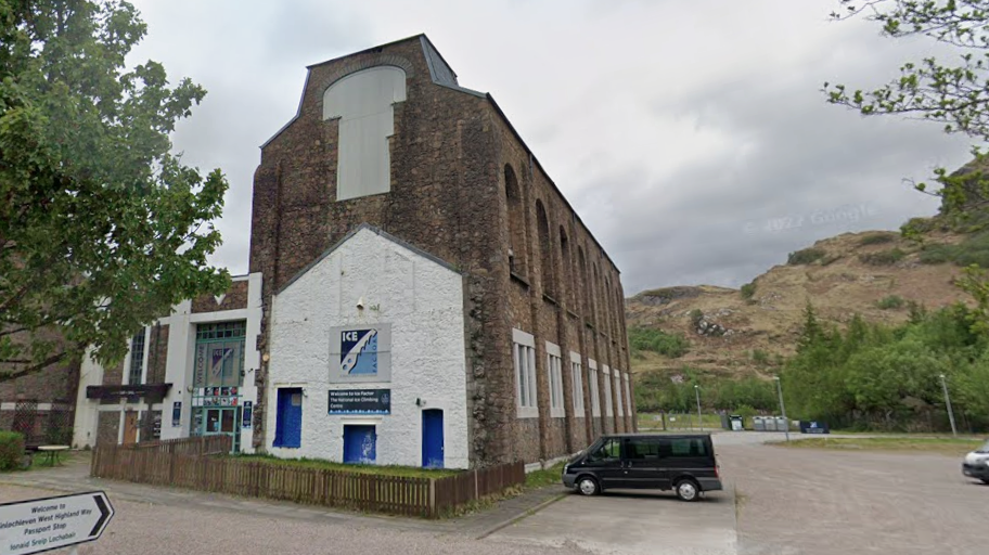 The former aluminium smelter, and Ice Factor premises, is tall, rectangular, red brick building. The view is of a gable end, and it has a number of blue doors. Next to the building is a large car park with a van parked in it. Behind the building are low, rugged hills.