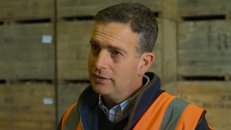 Robert Strathern, who has cropped dark hair, is standing in a potato cold store. He is wearing a high viz orange vest over a navy fleece and blue checked shirt. There are wooden boxes piled high behind him. 