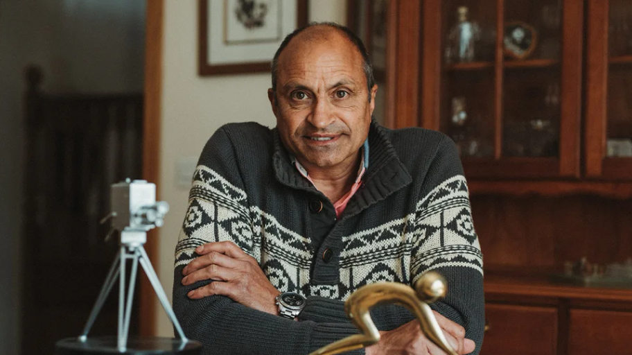 A man with short hair, wearing a grey and white patterned top, his arms folded. He is smiling at the camera