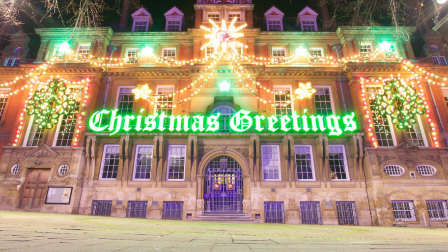 The town hall building adorned with lights and festooned with the illuminated banner saying Christmas Greetings