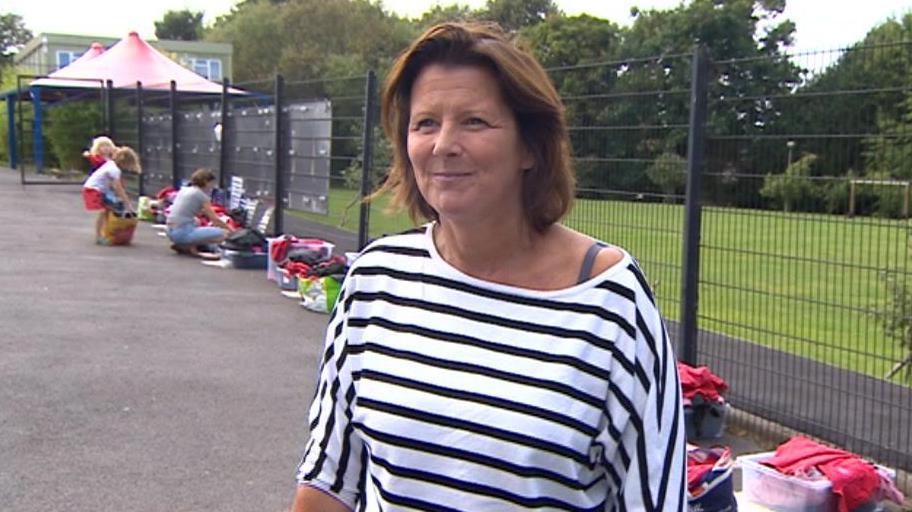 Amanda Pritchard standing in the school playground wearing a striped shirt. Behind her is a wire fence and lots of bags full of red school uniforms. A parent and two children are in the background browsing through the bags