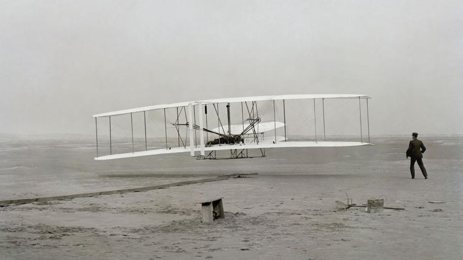 A black and white image of a plane used by the Wright brothers. A man stands to the left of the plane, with his back facing the camera.