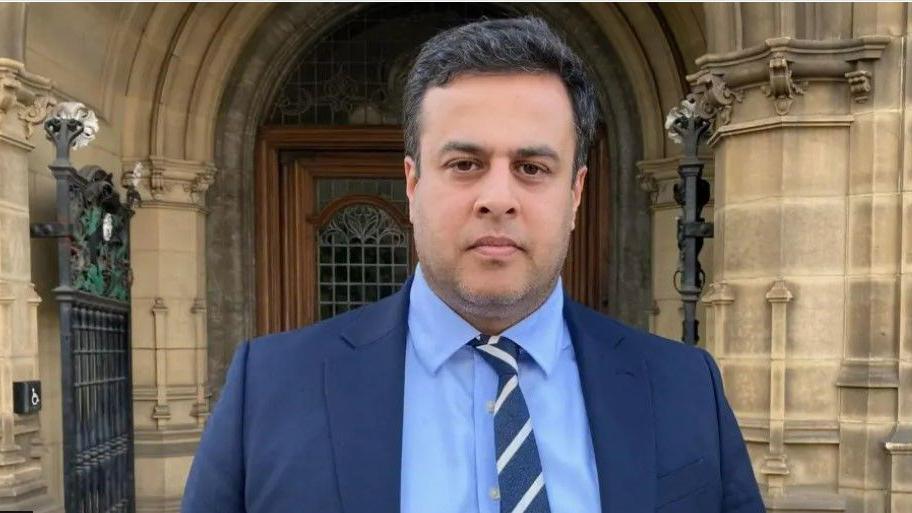 Nadeem Ahmed is wearing a blue suit with a pale blue shirt and blue and white striped tie and he stands outside the door of a stone-built building.