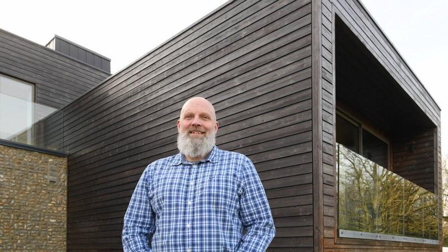 Man standing wearing a checked shirt with a painted black wooden panel wall of a house behind him - he has a white beard and is smiling into the camera